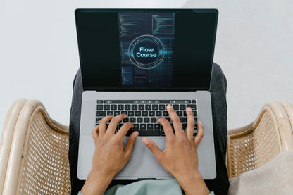 Top-down view of a person typing on a silver laptop with a black screen displaying 'Flow Course', seated in a woven chair.