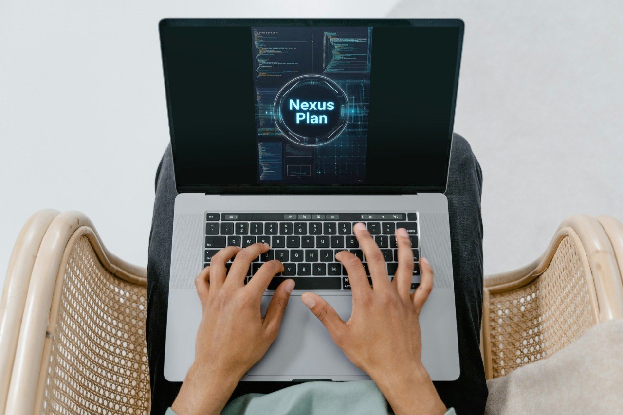 A top-down view of a person typing on a silver laptop with a black screen displaying 'Nexus Plan', seated between two woven beige armchairs.