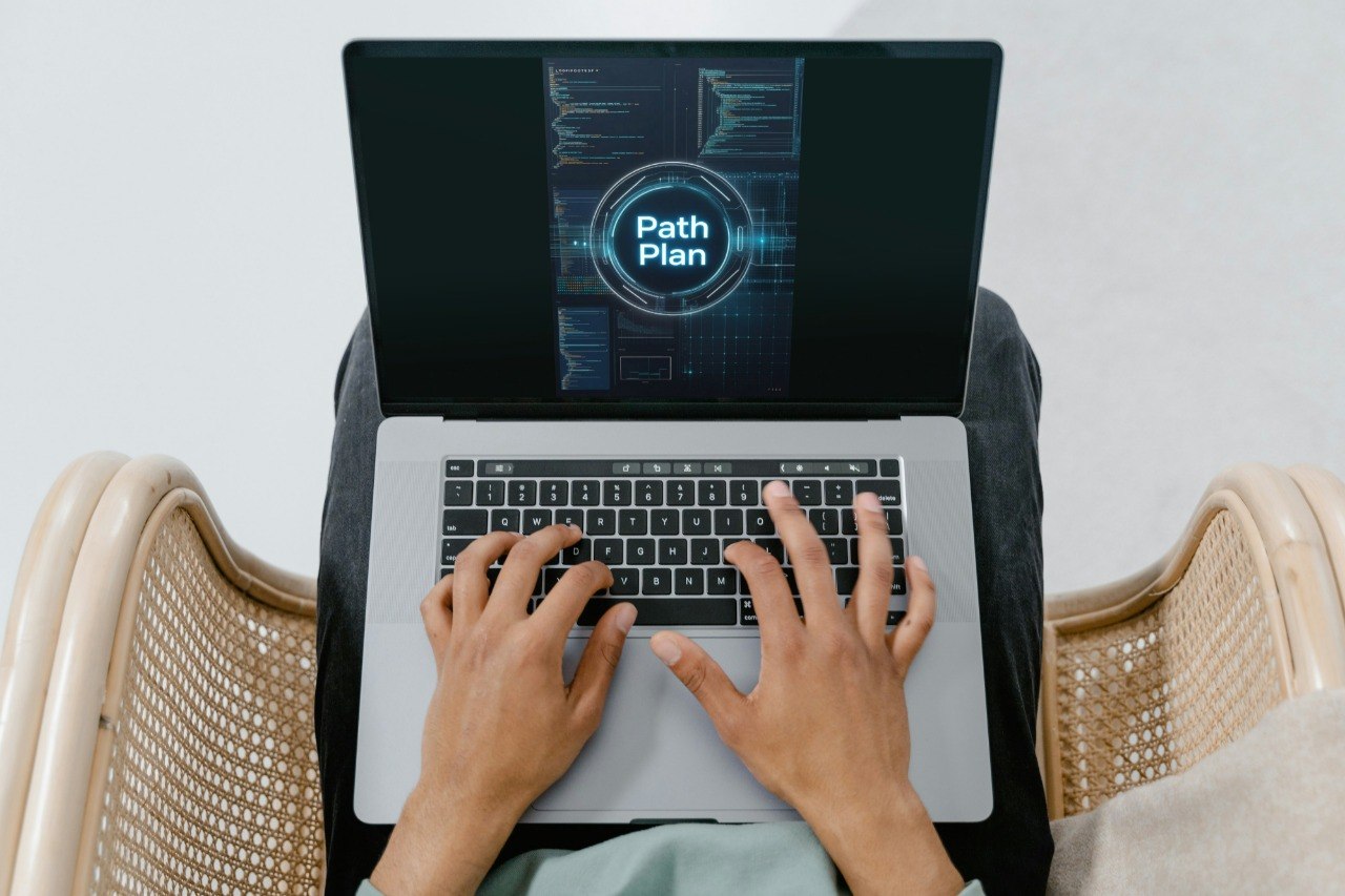 A top-down view of a person typing on a silver laptop with a glowing digital screen displaying 'Path Plan'. The person is seated in a woven cane chair against a light gray background.