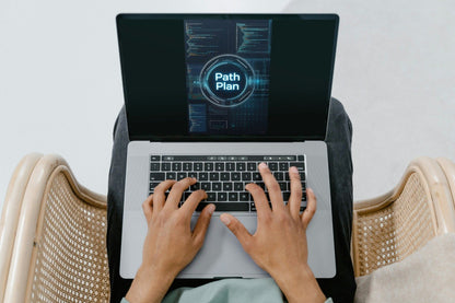 A top-down view of a person typing on a silver laptop with a glowing digital screen displaying 'Path Plan'. The person is seated in a woven cane chair against a light gray background.